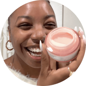 a dark skinned woman, @amanirakeia, smiling and holding jar of biossance squalane + vitamin c rose moisturizer